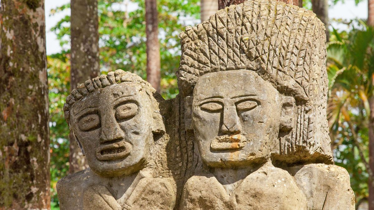 zur Vergrößerungsansicht des Bildes: Die Figuren einer Frau und eines Mannes im Stil der Mayas aus Stein mit großen Augen und wulstigen Lippen vor einigen tropischen Bäumen.